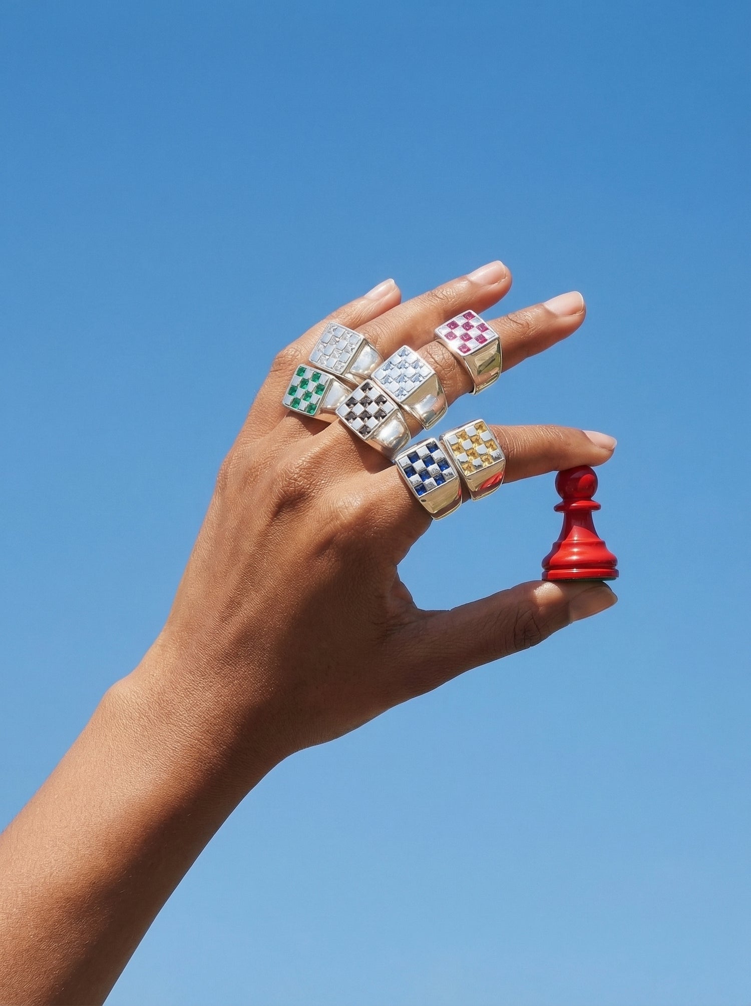 Hand wearing multiple colorful rings holding a red chess piece against a clear blue sky