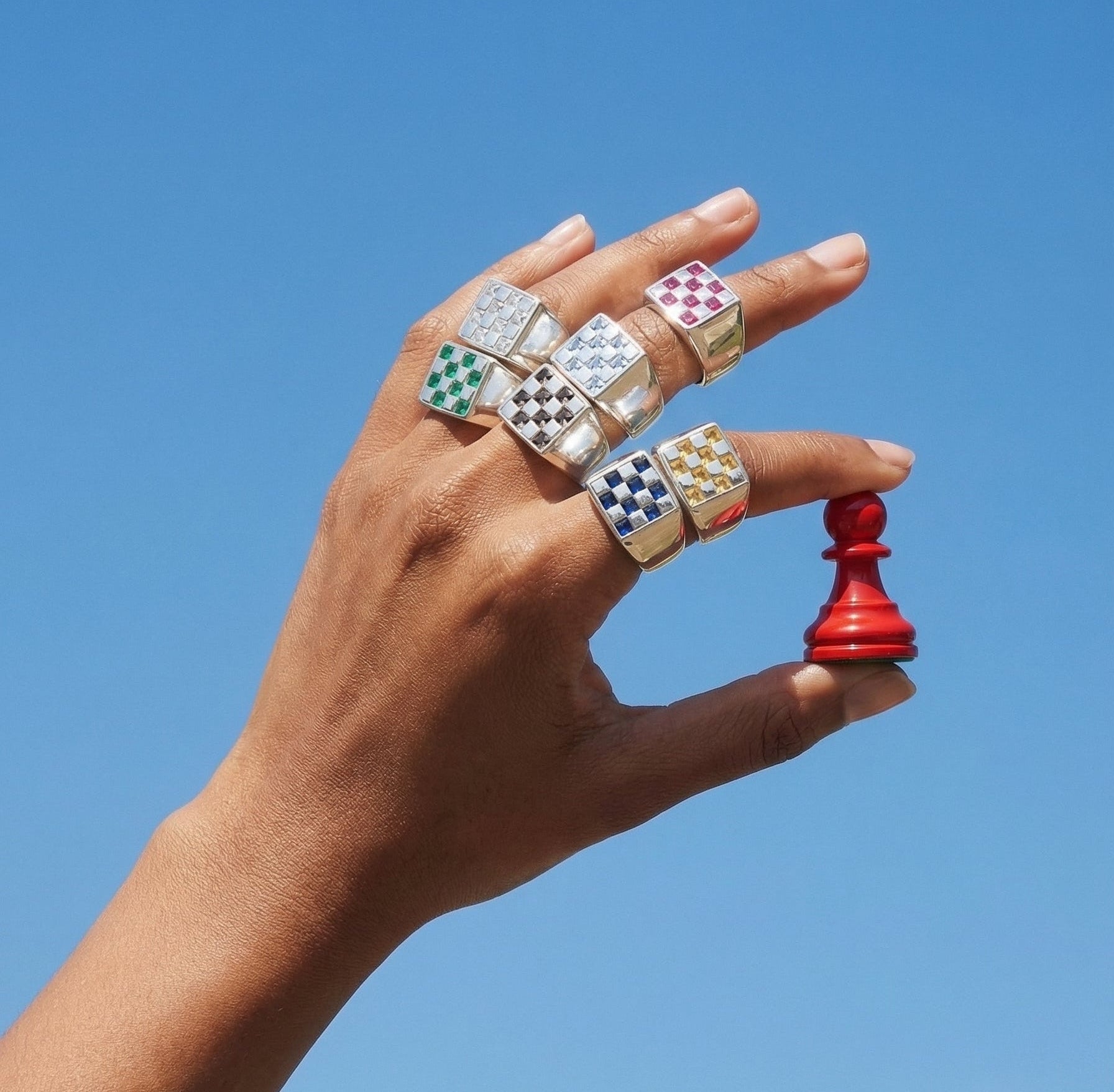 Hand wearing multiple colorful rings holding a red chess piece against a clear blue sky